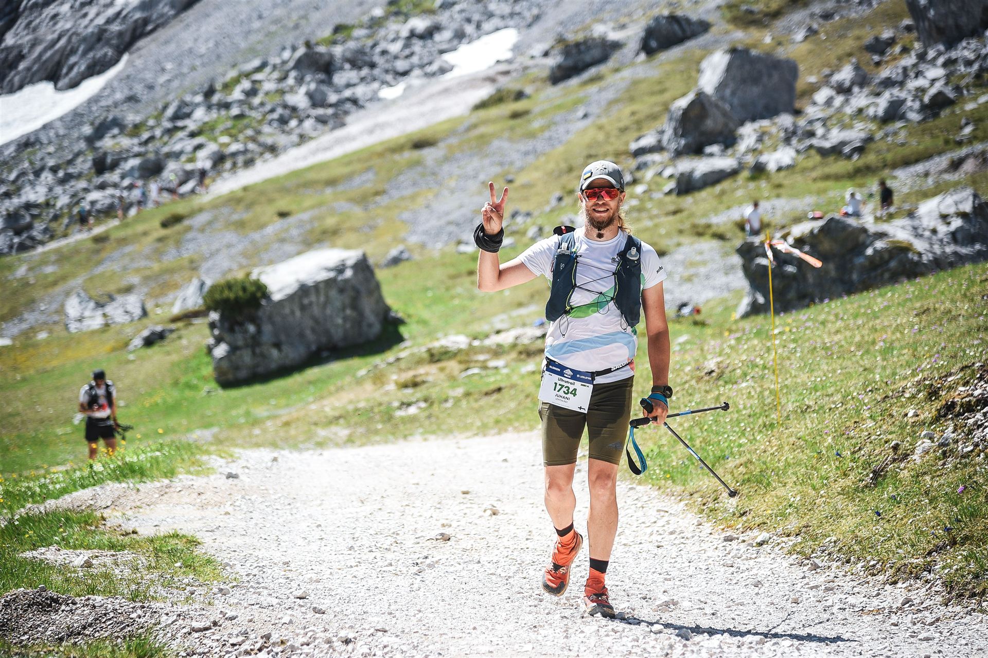 Juhani flashing a peace sign on a high alpine gravel road, mid-ultra, mountains behind